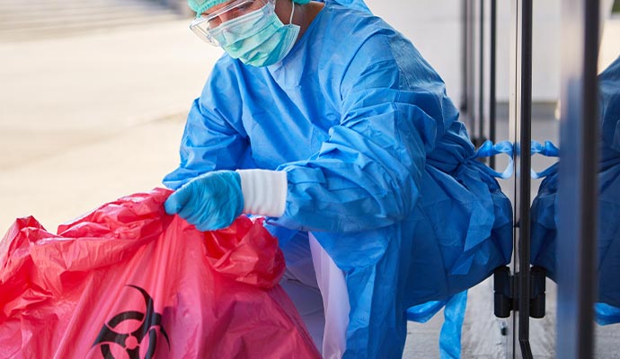 Worker in full safety gear handling red biohazard waste bag