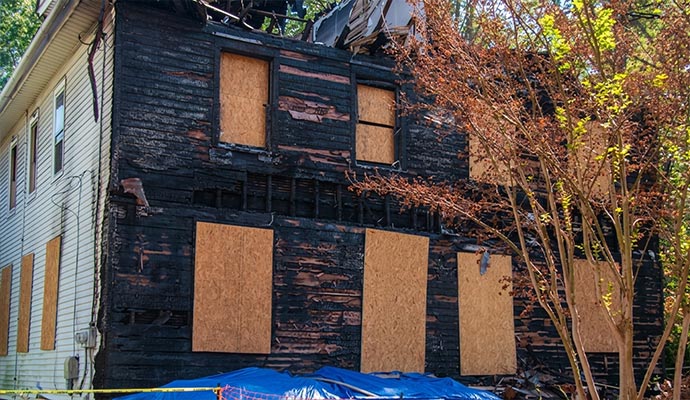 Fire-damaged house with windows boarded up and debris covered by tarp